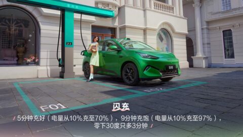 Green BYD Seagull plugged in at a charging station on a city street, with a woman standing beside the open driver's door and Chinese text listing the model's fast-charging times