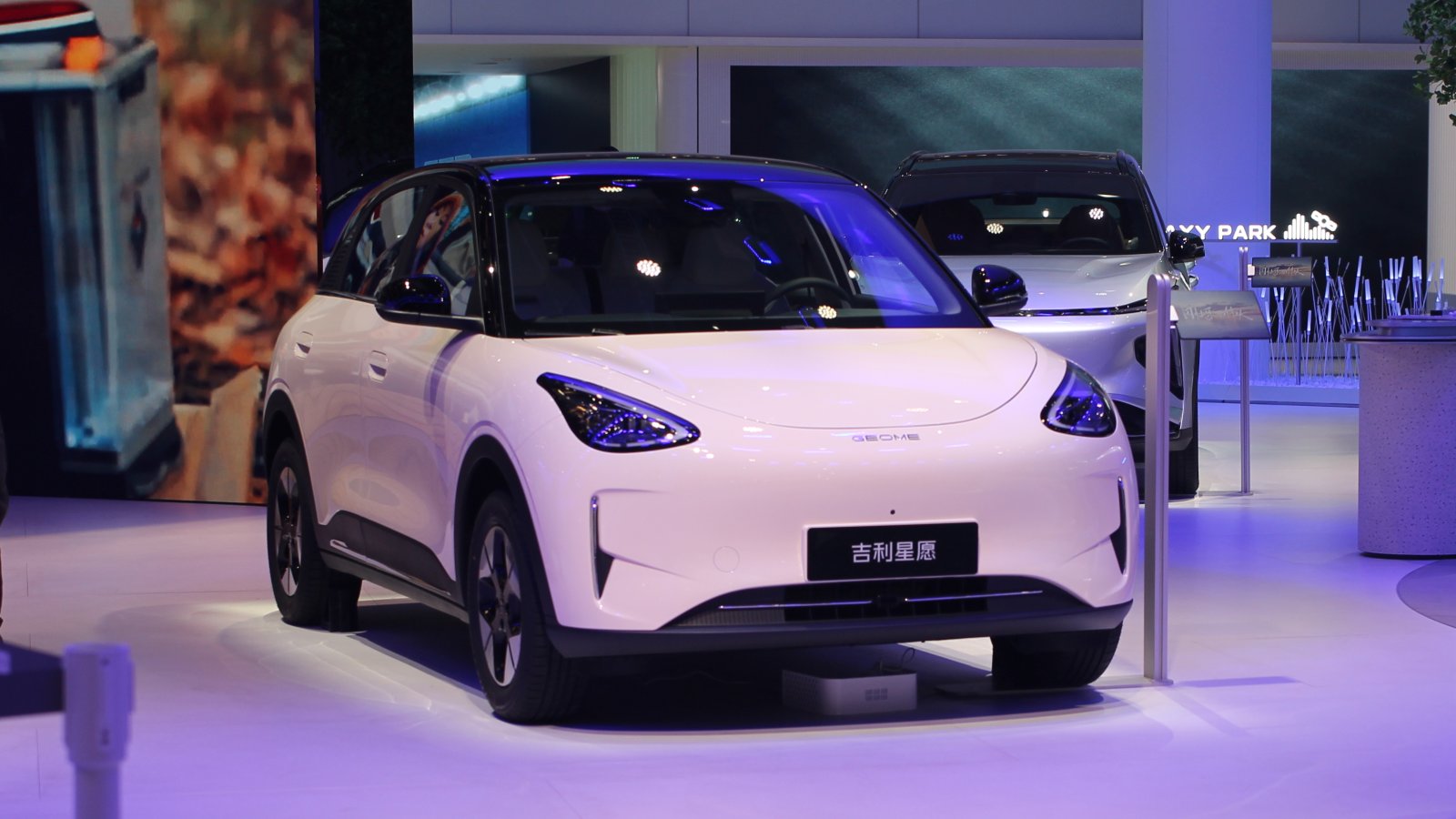A white Geely Xingyuan small electric hatchback with a black roof displayed on a stage at the Shanghai auto show, with a silver Geely Galaxy SUV in the background.