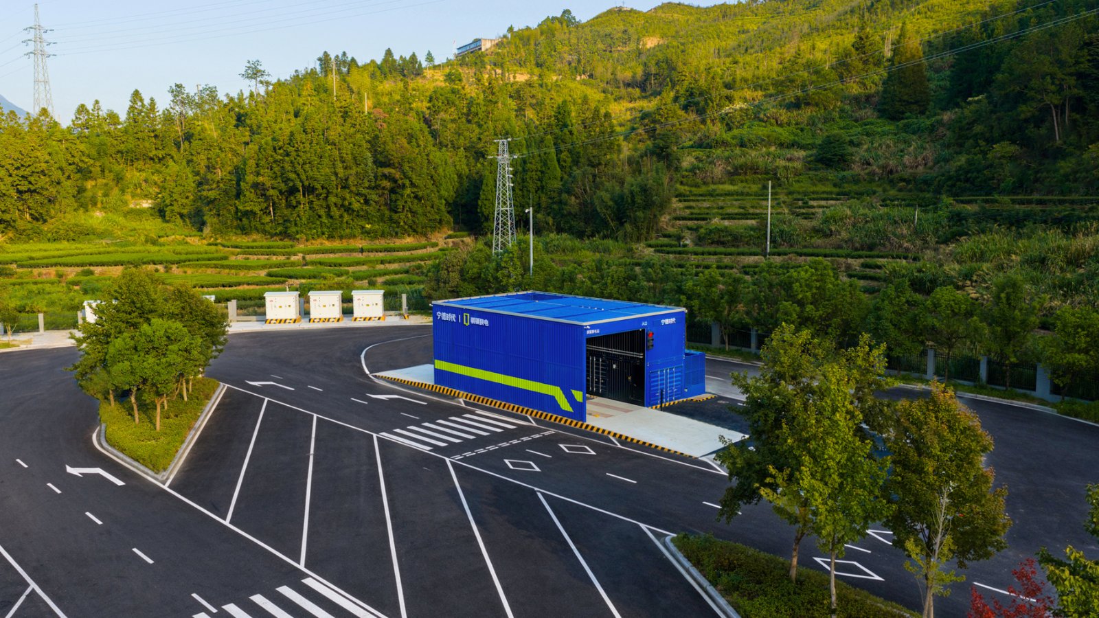 Aerial view of a blue CATL battery swap station for trucks, standing beside a newly paved road with terraced green hillsides and power lines behind it.
