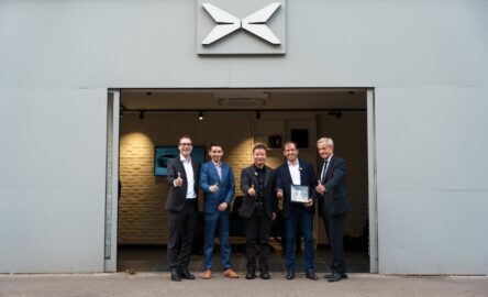 He Xiaopeng and 4 other men pose outside an Xpeng showroom, with 1 holding a presentation box.